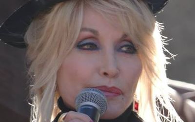 Dolly Parton, vestida con ropa de bombero, en Dollywood, presentando nuevas atracciones. Tiene un micrófono en sus manos.