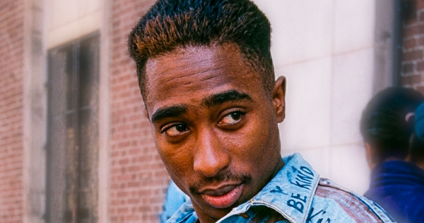 Image in QuizMyDay East Coast vs West Coast Hip Hop Trivia Quiz Challenge trivia quiz featuring 2Pac in 1988 on the cover of The Evening Sun, used by Baltimore Sun photographers as an example of modern school style, he is showing off a jeans jacket with Free Mandela written on the bottom, also you can see other hand written messages including hip hop written by the button holes with a mic drawn next to it. You can also see the peace sign on his chest pocket and above that it says Know what ⏱️ it is. You can also see The Born Busy Posse written on the side which was the name of his first rap group and on the collar it seems to say be kind, he also appears to be holding a cigarette